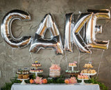 Decorative cake display with silver "CAKE" balloons and assorted desserts.